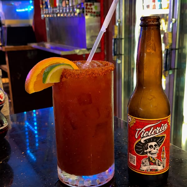 Spicy tomato cocktail with citrus garnish and straw beside a Victoria beer bottle on a colorful illuminated bar.