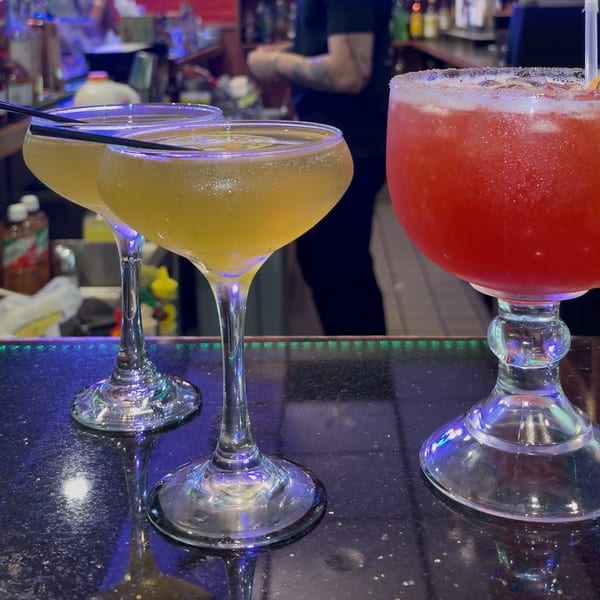 Three colorful cocktails sit on a reflective bar, including two pale yellow drinks and a red salted-rim beverage.