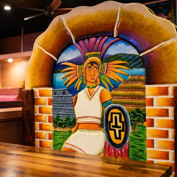 Restaurant booth with a colorful Aztec warrior painting on the backrest, and a TV in the background.