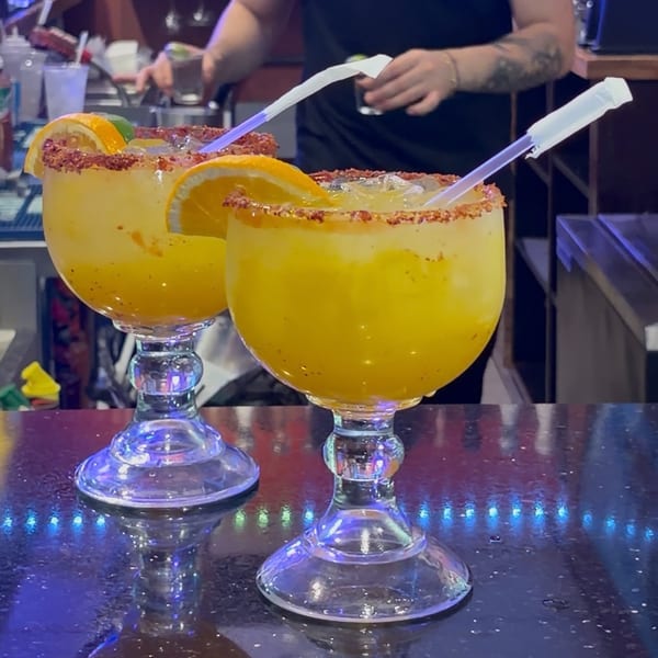 Two large orange cocktails with chili-salt rims, citrus garnishes, and straws sit on a brightly lit bar counter.